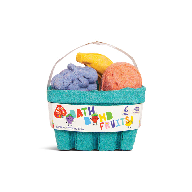 The Pacha Soap Co. Fruit Basket Bath Bomb Set features colorful, fruit-shaped bath bombs in a teal basket resembling grapes, a banana, and an orange. The label reads "Bath Bomb Fruits! 6 Pack" with playful fruit illustrations.