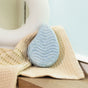 The Sand & Sea Bath Bomb by Pacha Soap Co., a blue teardrop shape with wavy grooves and clary sage, sits on a beige waffle towel on wood near a round white object and frosted glass window.