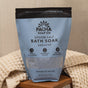 A bag of Pacha Soap Co. Breathe Salt Soak rests on a wooden surface beside a textured, cream-colored towel. Its blue and black packaging features crisp white text highlighting soothing Epsom salt and eucalyptus oil.
