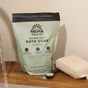 A green bag of Pacha Soap Co. Muscle Epsom Salt Soak, filled with mineral-rich Epsom and sea salts, sits on a wooden surface beside a folded towel and spoon as water pours in—ideal for relaxing tired muscles.