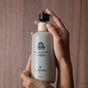 A person holds a Pacha Soap Co. Body Lotion bottle with one hand and presses the pump dispenser with the other. The lotion, infused with hydrating coconut oil, sits on a smooth wooden surface in the background.