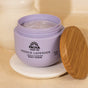 A lavender-hued jar of Pacha Soap Co. Body Scrub sits open on a beige surface, its wooden lid resting beside it. The whipped formula inside offers gentle exfoliation for smooth, refreshed skin.