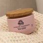 A pink jar of Pacha Soap Co. Body Scrub with nourishing oils and a wooden lid sits on a textured cream-colored blanket.