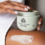 A person in a white towel applies Pacha Soap Co. Body Scrub to their leg, holding an open jar and spreading the nourishing exfoliating blend on their skin.