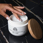A neatly manicured hand dips into a jar of Pacha Soap Co. Body Scrub on a dark marble surface, with the bamboo lid placed beside the open container.