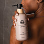 A person with brown skin holds a bottle of Pacha Soap Co. Body Wash against their wet shoulder, glistening water droplets on both the skin and the bottle.