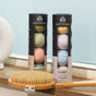 Two cylindrical packages of Pacha Soap Co.'s Froth Bomb and Pacha Peaces bath bombs sit on a wooden shelf next to a dry body brush. Each is filled with vibrant bath bombs, while a white object is partially visible in the background.