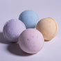 Four pastel Froth Bomb bath bombs (lavender, pink, blue, yellow) by Pacha Soap Co. are neatly arranged on a light surface as part of the Pacha Peaces set, casting soft shadows.