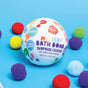 A colorful Mystery Bath Bomb by Pacha Soap Co. sits on a blue surface with bright pom-poms, featuring playful packaging with question marks and teasing a surprise color and fruity scent inside.