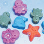 Bar Soap Buddies by Pacha Soap Co. are colorful sea animal bath toys—turtles, octopuses, a starfish, and a dolphin—each with a smiling face. They rest on a blue surface with gentle soap bubbles.