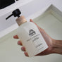 A hand holds a bottle of Pacha Soap Co. Liquid Hand Soap with a black pump and wooden collar over a white sink.