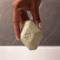 A hand with manicured nails holds the Pacha Soap Co. Muscle Epsom Salt Bath Bomb, a light green hexagonal bath bomb with a leaf imprint, ideal for aromatherapy and soothing muscles, against a blurred dark background.