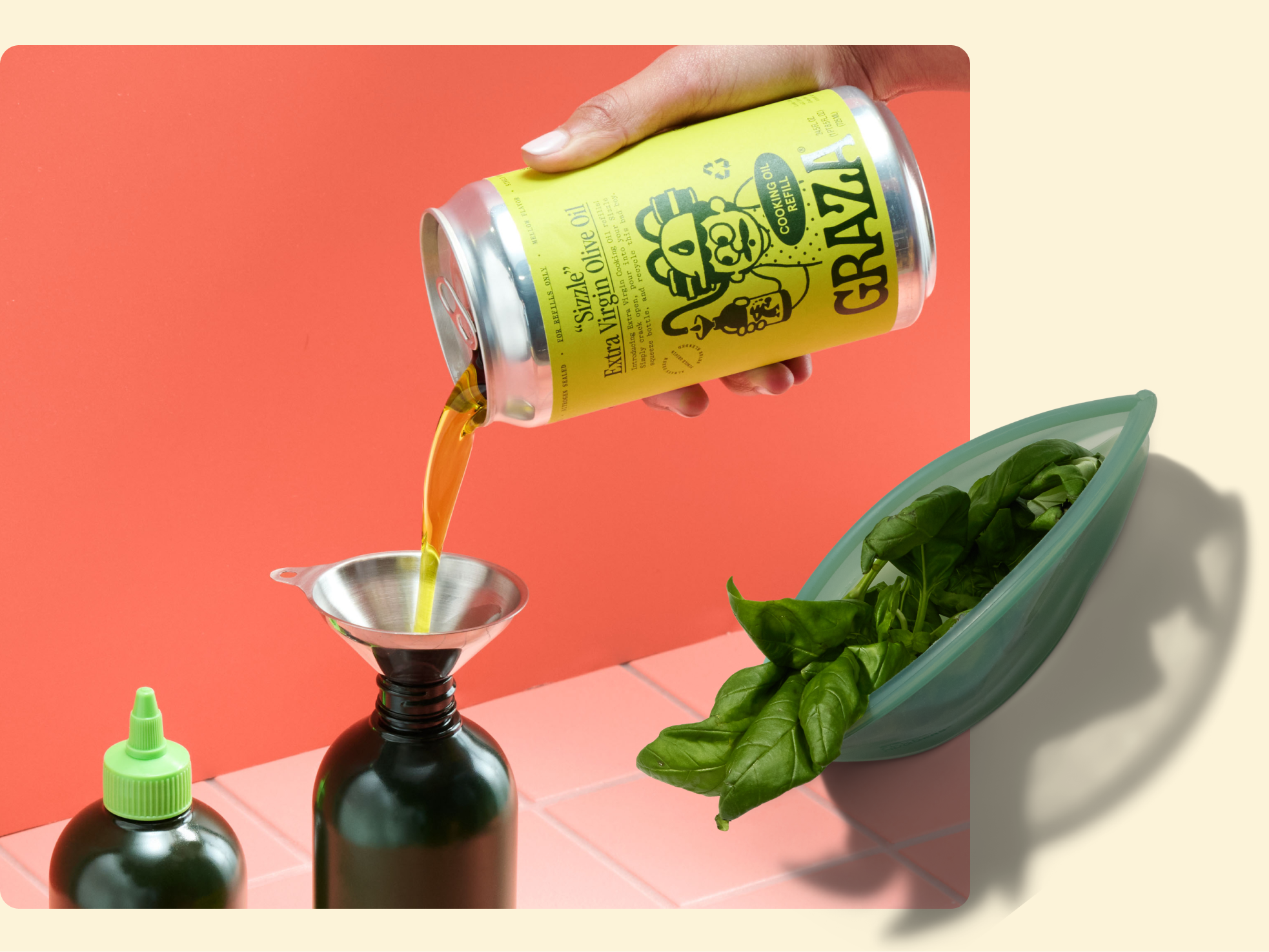 A hand pours Graza extra virgin olive oil from a can into a dark glass bottle with a funnel, next to a green bowl filled with fresh basil leaves on a pink tiled surface against a coral background.