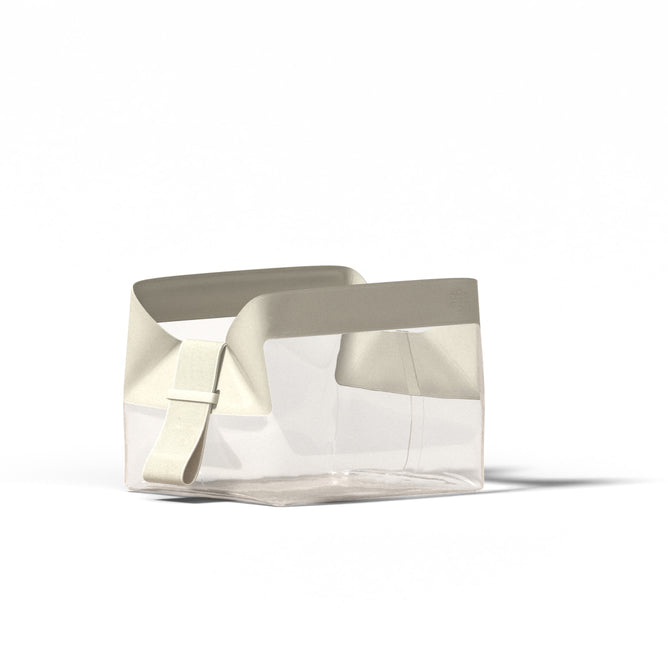 The Parcel by Cadence is a transparent rectangular TSA-approved travel bag with wide off-white handles, an adjustable strap, and a magnetic travel system—shown against a plain white background.