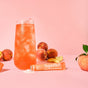 A tall glass of iced peach-colored drink sits among fresh peaches and two orange blume SuperBelly Probiotic Hydration Powder packets on a pink background.