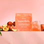A peach-colored packet of blume SuperBelly Probiotic Hydration Powder sits beside a glass of matching drink and fresh peaches, set against a soft pink background.