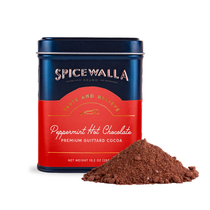 A tin labeled Deluxe Hot Chocolate - Peppermint by Spicewalla sits behind cocoa powder. The red and blue 10.2 oz tin features premium Guittard cocoa—perfect for a festive holiday hot chocolate treat.