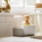 A white soap dish holds a scrub brush with a wooden handle and a Full Circle Plain Jane - Cellulose Sponge, set on a kitchen counter near the sink and glass soap dispenser. Sunlight streams in through the window above.