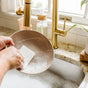 Washing a soapy beige plate at the kitchen sink, someone uses a Full Circle Plain Jane - Cellulose Sponge. Gold faucets, a soap dispenser, and sunlight streaming through the window complete the scene.