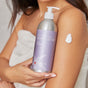A person wrapped in a towel holds Plaine Products Body Lotion, Citrus Lavender, against their arm and applies a dab to their upper arm. The label features the scent “Citrus Lavender” and mentions soothing organic aloe vera.