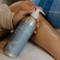 A person applies Plaine Products Body Lotion, Rosemary Mint Vanilla, from a silver pump bottle to their leg while seated on a bed with a blue and white blanket. The nourishing formula features organic aloe vera.