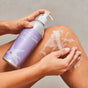 A person applies Plaine Products Body Wash, Citrus Lavender onto their leg from a silver and purple pump bottle, creating a wet, lathered layer of this skin-soothing soap.