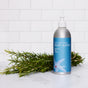 A silver Plaine Products Body Wash, Rosemary Mint Vanilla bottle with a blue label sits on a white surface near fresh rosemary sprigs, emphasizing its natural ingredients, against a white subway tile background.