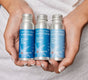 A person with manicured nails holds three small silver Plaine Products bottles—shampoo, conditioner, and body wash—all in the Rosemary Mint Vanilla scent, made with natural ingredients for a zero waste routine.