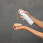 A person dispenses Plaine Products Body Wash, Unscented from a silver bottle onto a round wooden bath brush with light bristles, set against a plain gray background.