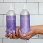 Two hands hold Plaine Products Conditioner, Citrus Lavender bottles with pump tops under running water. The vegan formula hydrates hair, featuring refreshing citrus and soothing lavender scents, set against a white tiled background.