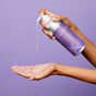 A person dispenses Plaine Products Conditioner, Citrus Lavender from a purple can onto their palm with a plain lavender background.