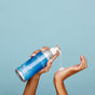A person pumps Plaine Products’ Rosemary Mint Vanilla vegan conditioner from a silver and blue bottle onto their palm, promoting scalp balance against a plain light blue background.