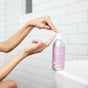 In a white-tiled bathroom with a glimpse of a bathtub, someone pumps Plaine Products Deep Conditioner onto their hand to restore shine to their hair.