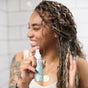 A smiling person with braided hair sprays Plaine Products Hair Repair detangler in a tiled bathroom, raising one hand near their face.