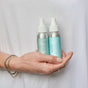 A person in a white shirt and gold bracelets holds two Plaine Products silver bottles—Hair Repair and Sea Salt Spray—hair repair and detangler products with blue-green labels.