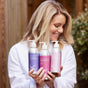 A woman in a white robe smiles while holding Plaine Products Purple Shampoo and other vegan, cruelty-free hair care items against a blurred outdoor background.