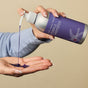A person dispenses Plaine Products Purple Shampoo, a vegan and cruelty-free formula that neutralizes brassy hair, from a sleek silver and purple bottle onto their palm against a beige background.