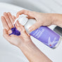 A person pumps Plaine Products Purple Shampoo from a silver and purple bottle onto their palm over a white sink, ready to neutralize brassy tones with this vegan hair care essential.