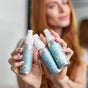 A person with long red hair holds up Plaine Products’ Sea Salt Spray, Hair Repair, and Styling Gel—three silver essentials for a beach-inspired look—with a blurred background.