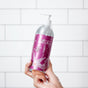 A hand holds a silver and pink Plaine Products Shampoo, Cherry Almond bottle with soap suds against white tiles. This vegan formula helps reduce frizz for smooth, nourished hair.