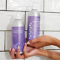 Two hands hold two Plaine Products Citrus Lavender vegan shampoo bottles with pump dispensers against a white tiled wall. One bottle displays the front label, the other shows back instructions—both promise hydrated hair.