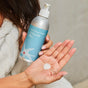 A person in a white robe holds a silver bottle of Plaine Products Styling Gel and dispenses some onto the palm of their other hand.