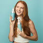 A smiling woman with long red hair holds a bottle of Plaine Products Styling Gel against a light blue background, showcasing her favorite vegan hair product.