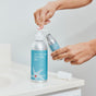 Someone dispenses Plaine Products Styling Gel from a silver pump bottle into a smaller refill bottle over the bathroom sink.