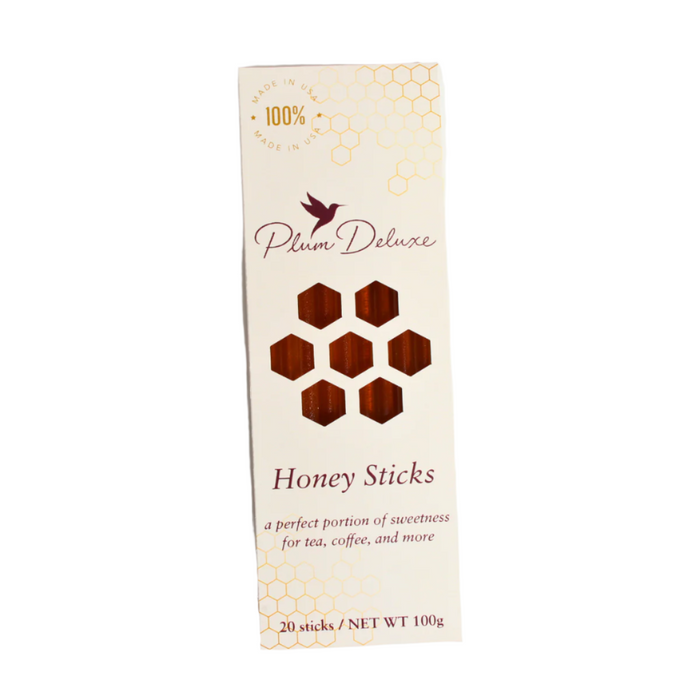 Plum Deluxe Honey Sticks come in a cream-colored box with gold accents, honeycomb patterns, and a bird logo. The packaging features honeycomb cutouts that reveal the tasty sticks inside—perfect for pairing with tea or wildflowers.