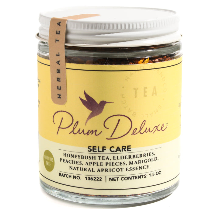 A glass jar of Plum Deluxe Self Care Apricot Elderberry Tea with a yellow label lists ingredients like honeybush blend, elderberry, peaches, apple pieces, and marigold. The jar has a white lid and a beige tag reading Herbal Tea.