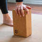 A person’s hand rests on the Yoloha Yoga Cork Yoga Block, which is placed on a mat with bare feet visible in the background. The block features a small flower-like logo on its surface, highlighting both its stylish design and supportive function for yoga practice.