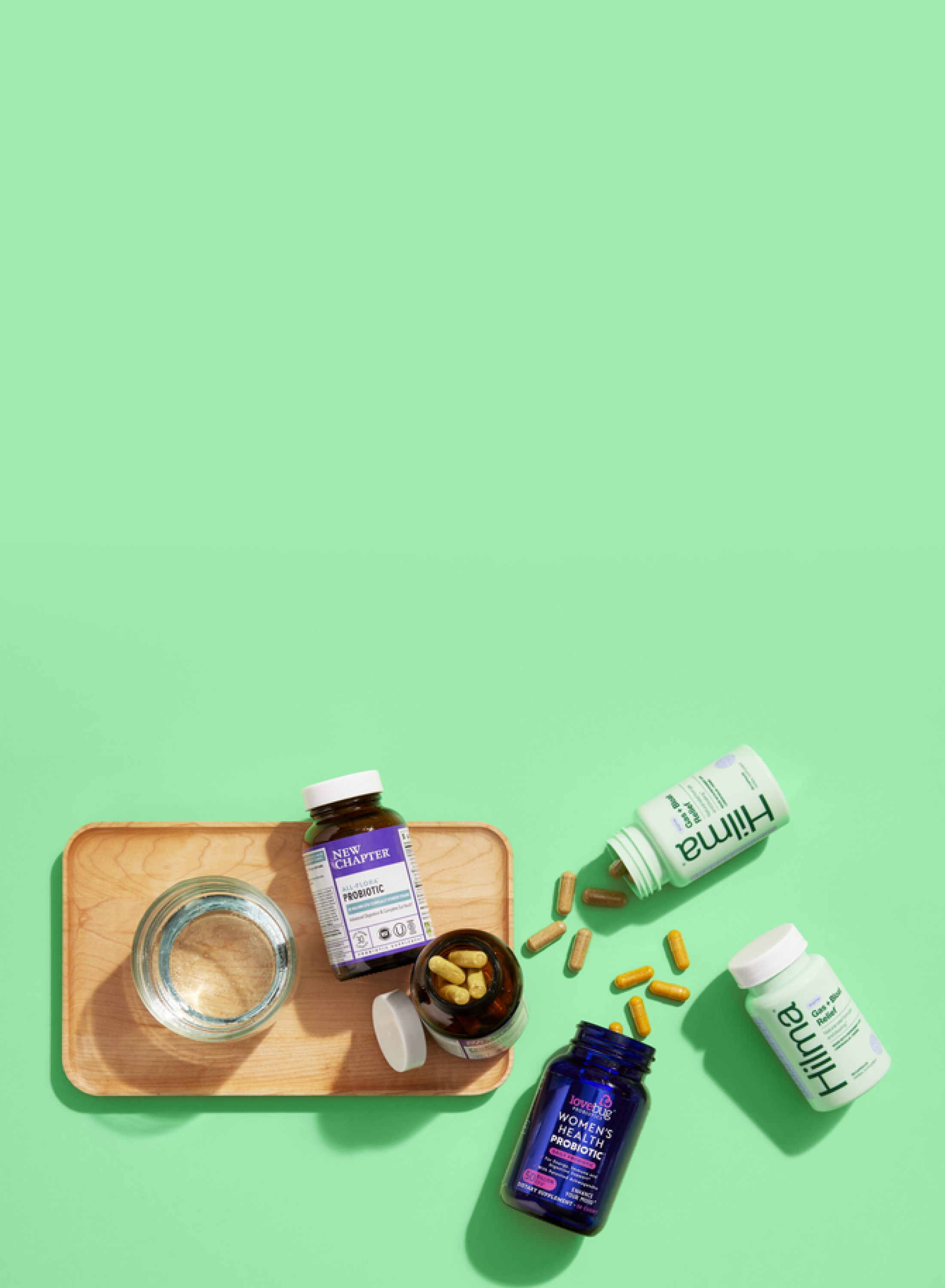 Supplements in green and blue bottles, some capsules scattered, on a green surface. A wooden tray with a glass of water and an open supplement bottle sits to the left. The background is a solid light green.