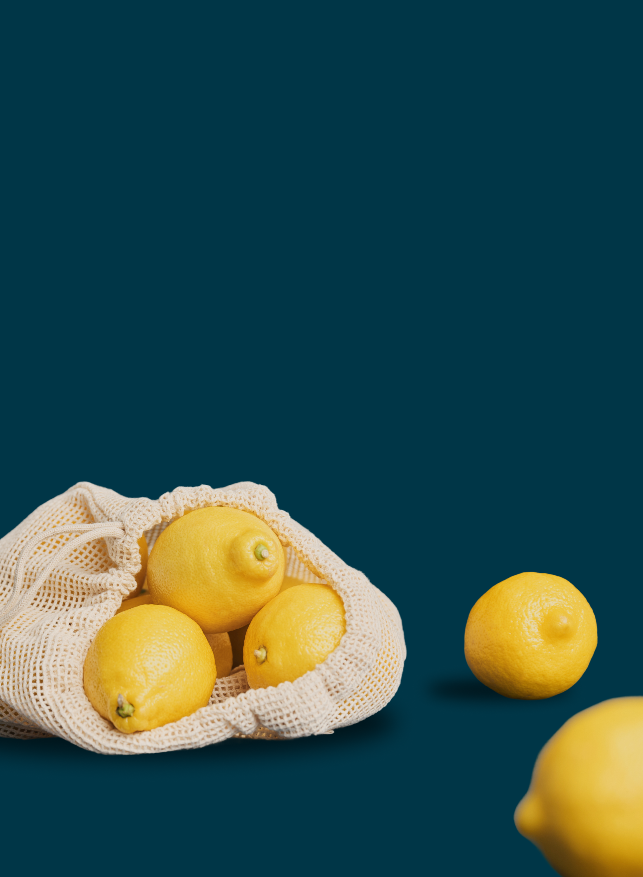 Fresh yellow lemons spilling out of an open cream cotton mesh reusable produce bag, with two loose lemons nearby, against a deep teal background.