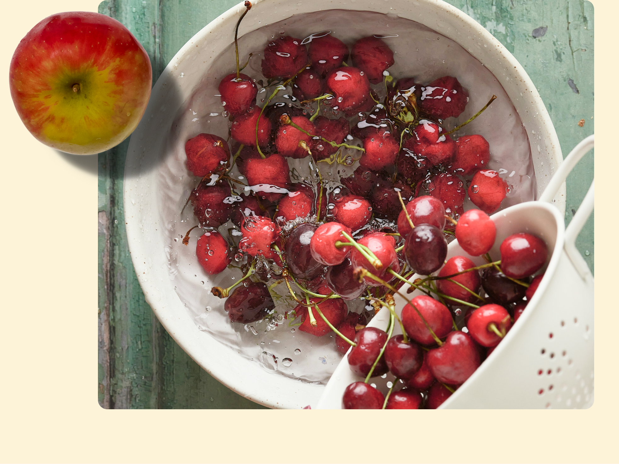 A bowl of cherries being washed with water, some cherries in a white colander, and a single apple placed on a green wooden surface.